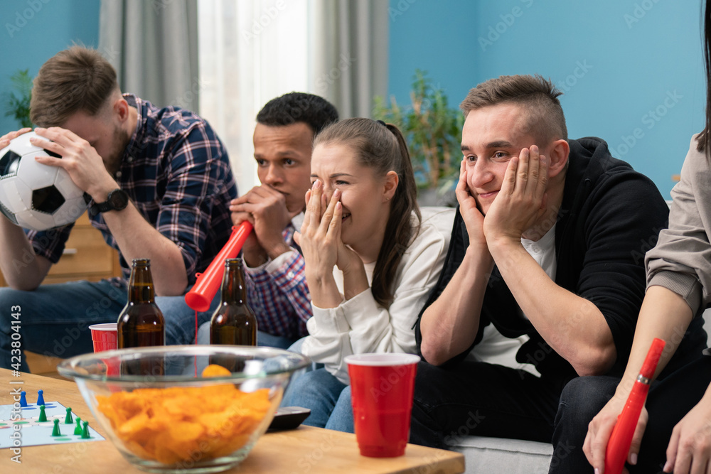 Upset friends watching football and drinking at home sitting on a couch. Students watching sport on tv together at home team lost. Angry young friends are frustrated Upset friends watching football and drinking at home sitting on a couch. Students watching sport on tv together at home team lost. Angry young friends are frustrated