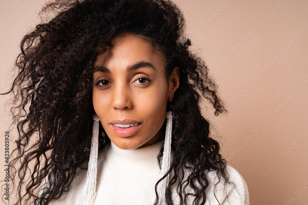Portrait of dark-skinned woman with curly hairs smiling on beige backdrop. Looking into camera . Wearing stylish earrings and knitted sweater. Portrait of dark-skinned woman with curly hairs smiling on beige backdrop. Looking into camera . Wearing stylish earrings and knitted sweater.