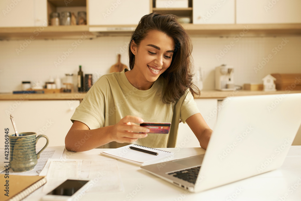 Stylish pretty student girl surfing internet in kitchen, sitting at table with open laptop and mug, holding plastic credit card, entering information via paying system while shopping online - obrazy, fototapety, plakaty 