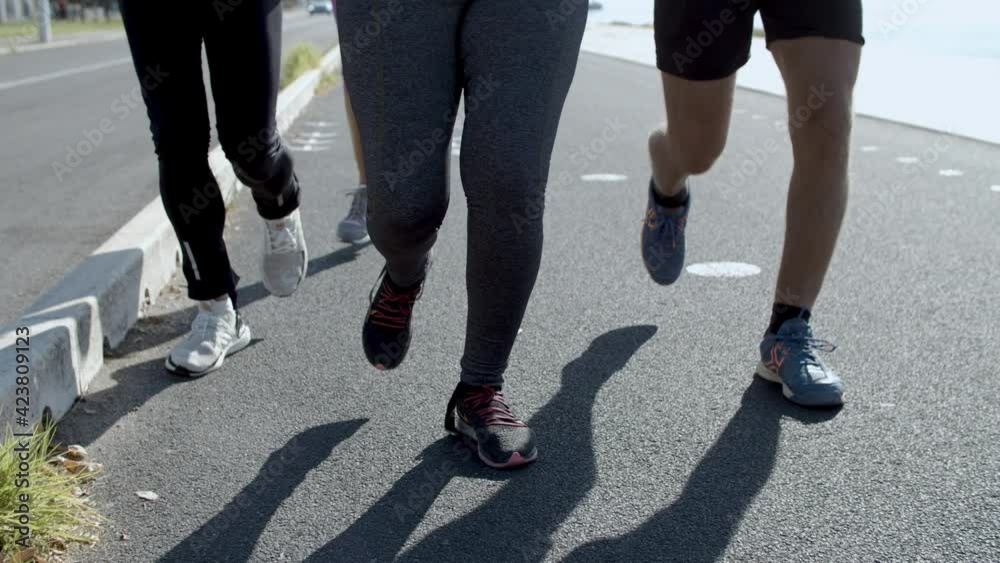 Sporty confident runners exercising together outdoors in sunny day. Closeup human legs jogging on asphalt road. Vertical motion. Tracking camera. Healthy lifestyle and sport activity concept Sporty confident runners exercising together outdoors in sunny day. Closeup human legs jogging on asphalt road. Vertical motion. Tracking camera. Healthy lifestyle and sport activity concept