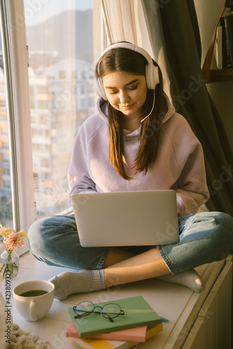 Smiling woman student in headphone study online with laptop at home. Girl sitting near window on sunset. Distance learning and online teaching concept.