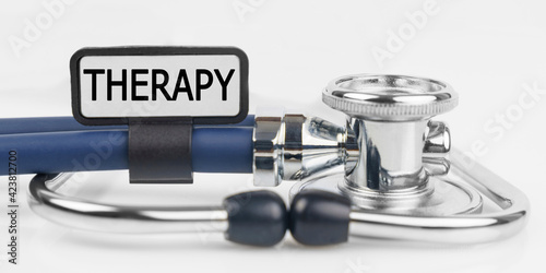 On the white surface lies a stethoscope with a plate with the inscription - THERAPY