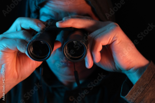 middle-aged man, stalker in hood looks through black binoculars in dark, peeps, illegally tracks down personal secrets of neighbors, concept of industrial espionage, surveillance