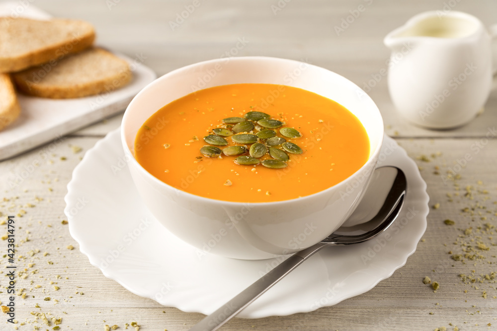 Homemade pumpkin and carrot soup in a white bowl with cream and seeds close up on white wooden rustic background Homemade pumpkin and carrot soup in a white bowl with cream and seeds close up on white wooden rustic background