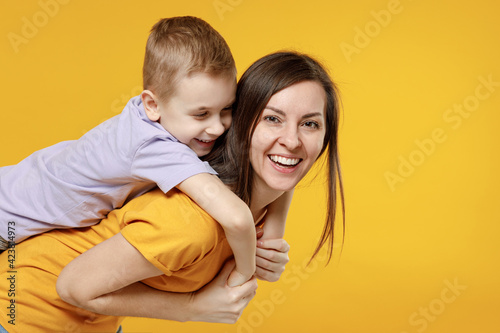 Happy young woman have fun with cute child baby boy 5-6-7 years old in violet t-shirt stand behind hug kiss Mommy little kid son together isolated on yellow background studio Mother's Day love family