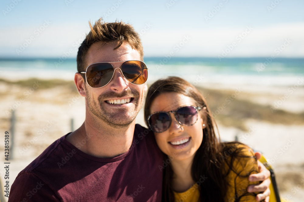 Portrait of happy caucasian couple sitting on beach by the sea embracing wearing sunglasses smiling