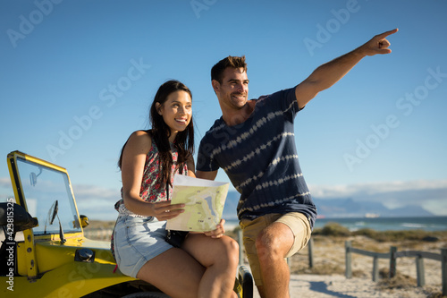 Happy caucasian couple sitting on beach buggy by the sea reading roadmap pointing
