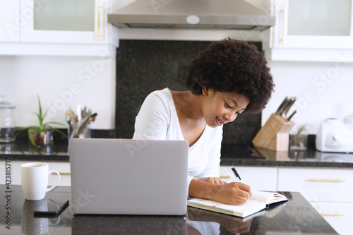 African american woman sitting in kitchen using laptop and taking notes