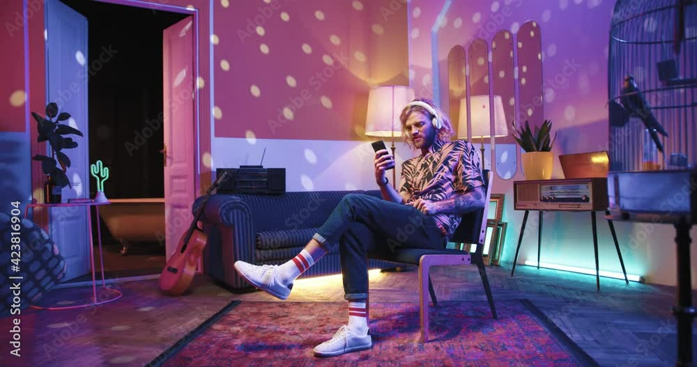 Young stylish Caucasian cool man sit on chair lin vintage apartment listening to music in headphones at home in room with warm light and typing on cellphone searching internet surfing social network