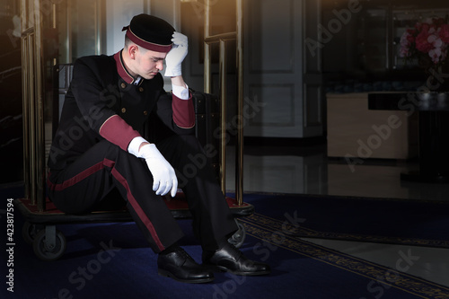 A tired doorman sits on a luggage cart. Uniformed doorman with white gloves. A young man. Hotel business. Hard work.Free space. photo