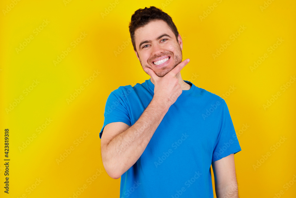 young handsome caucasian man wearing blue t-shirt against yellow background looking confident at the camera smiling with crossed arms and hand raised on chin. Thinking positive. young handsome caucasian man wearing blue t-shirt against yellow background looking confident at the camera smiling with crossed arms and hand raised on chin. Thinking positive.