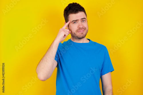 young handsome caucasian man wearing blue t-shirt against yellow background tries to memorize something, keeps fore finger on temple, reminds information for exam,