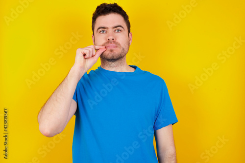 young handsome caucasian man wearing blue t-shirt against yellow background mouth and lips shut as zip with fingers. Secret and silent, taboo talking.