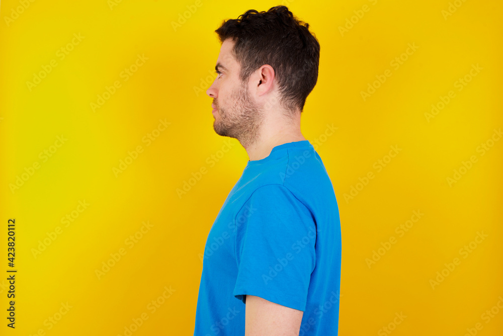 young handsome caucasian man wearing blue t-shirt against yellow background looking to side, relax profile pose with natural face with confident smile. young handsome caucasian man wearing blue t-shirt against yellow background looking to side, relax profile pose with natural face with confident smile.