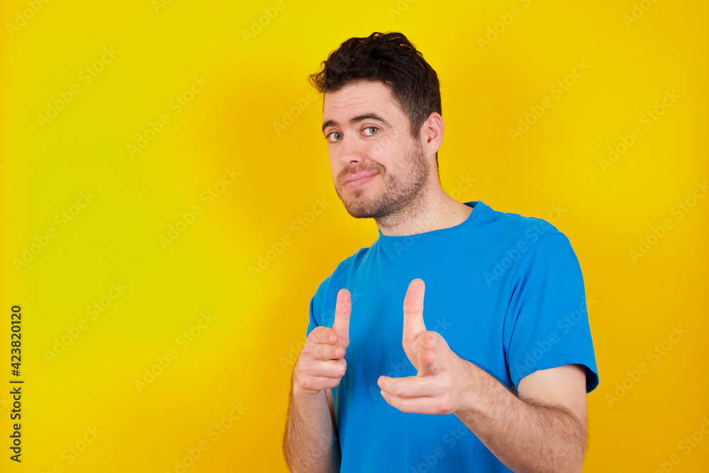 Joyful young handsome caucasian man wearing blue t-shirt against yellow background wink and points index fingers at camera, chooses someone, makes finger gun pistol. Joyful young handsome caucasian man wearing blue t-shirt against yellow background wink and points index fingers at camera, chooses someone, makes finger gun pistol.