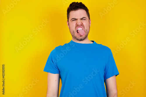 young handsome caucasian man wearing blue t-shirt against yellow background sticking tongue out happy with funny expression. Emotion concept.
