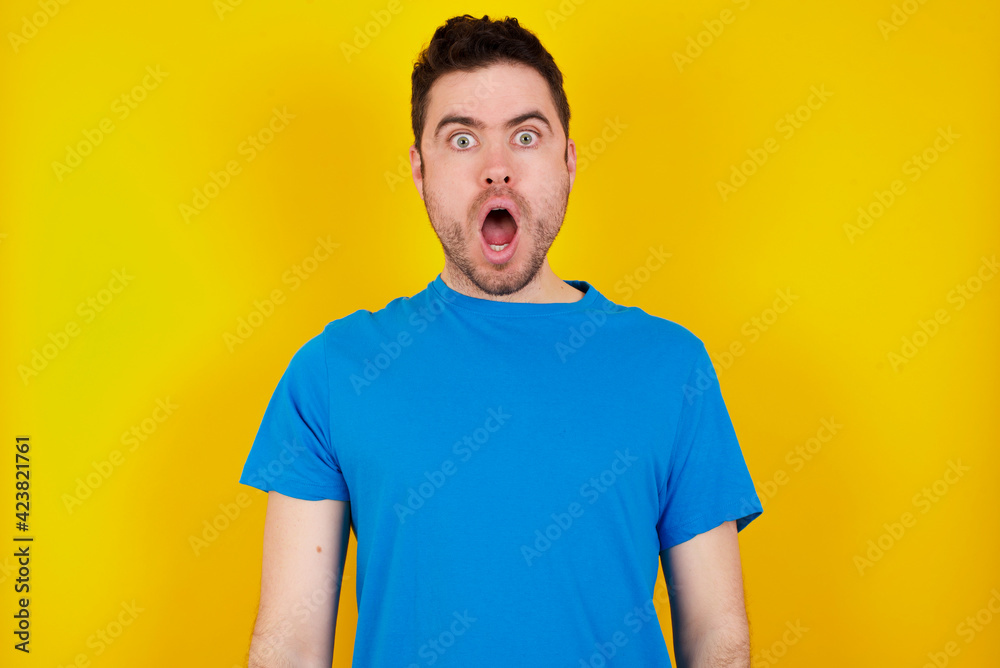 Frustrated young handsome caucasian man wearing blue t-shirt against yellow background feels puzzled and hesitant, shrugs shoulders in bewilderment, keeps mouth widely opened, doesn't know what to do. Frustrated young handsome caucasian man wearing blue t-shirt against yellow background feels puzzled and hesitant, shrugs shoulders in bewilderment, keeps mouth widely opened, doesn't know what to do.
