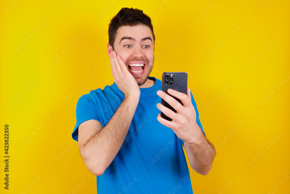 Portrait of young handsome caucasian man wearing blue t-shirt against yellow background holding in hands cell celebrating Portrait of young handsome caucasian man wearing blue t-shirt against yellow background holding in hands cell celebrating