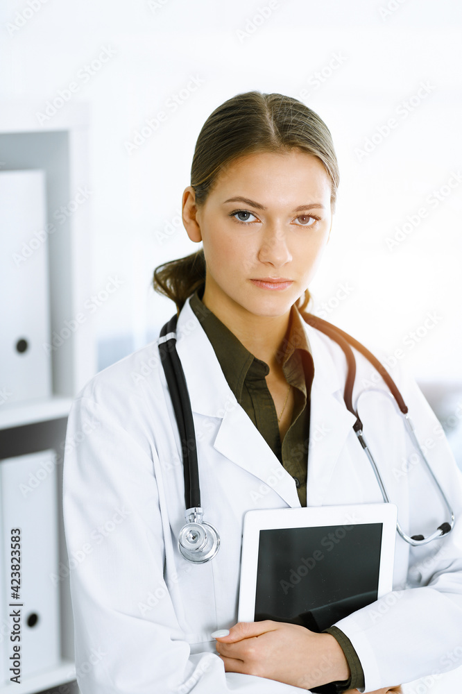 Woman-doctor standing and looking at camera in sunny clinic. Medicine and healthcare Woman-doctor standing and looking at camera in sunny clinic. Medicine and healthcare