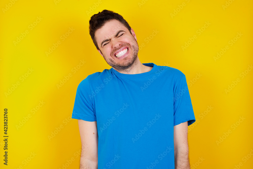 Positive young handsome caucasian man wearing blue t-shirt against yellow background with overjoyed expression closes eyes and laughs shows white perfect teeth Positive young handsome caucasian man wearing blue t-shirt against yellow background with overjoyed expression closes eyes and laughs shows white perfect teeth