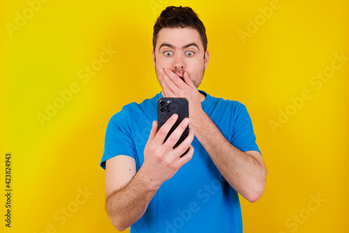 young handsome caucasian man wearing blue t-shirt against yellow background being deeply surprised, stares at smartphone display, reads shocking news on website, Omg, its horrible!