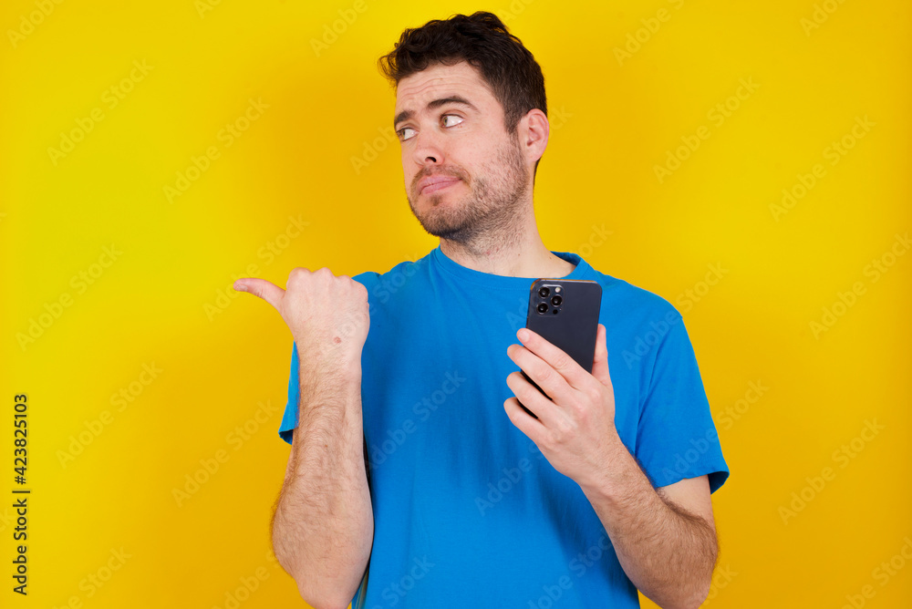 young handsome caucasian man wearing blue t-shirt against yellow background points thumb away and shows blank space aside, holds mobile phone for sending text messages. young handsome caucasian man wearing blue t-shirt against yellow background points thumb away and shows blank space aside, holds mobile phone for sending text messages.
