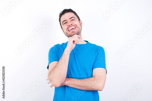 Optimistic young handsome caucasian man wearing blue t-shirt against white background keeps hands partly crossed and hand under chin, looks at camera with pleasure. Happy emotions concept.