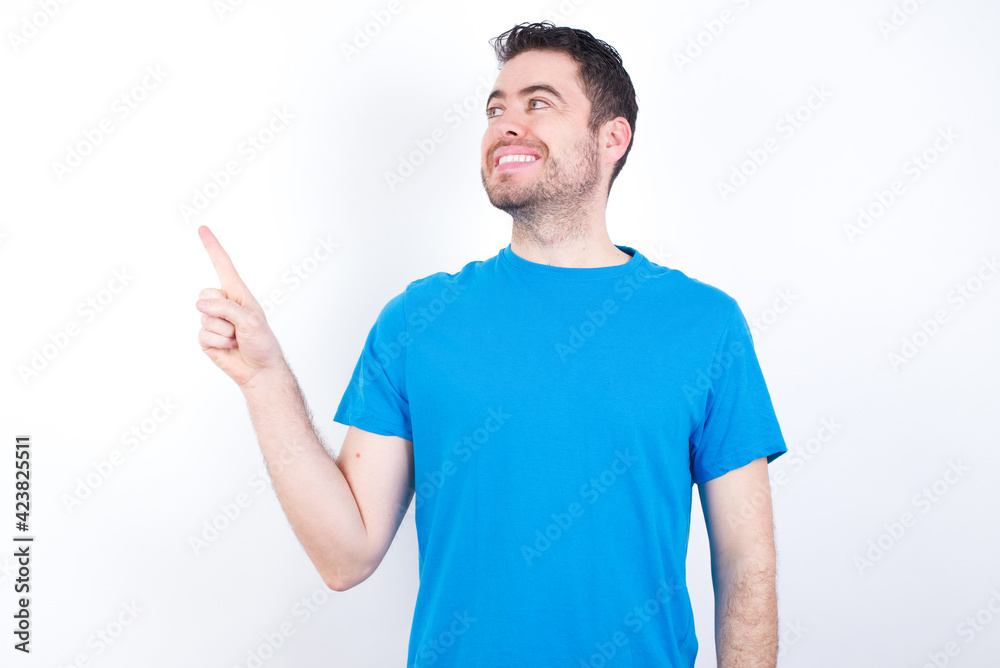 Smiling young handsome caucasian man wearing blue t-shirt against white background indicating finger empty space showing best low prices Smiling young handsome caucasian man wearing blue t-shirt against white background indicating finger empty space showing best low prices