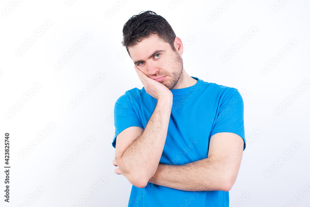 Very bored young handsome caucasian man wearing blue t-shirt against white background holding hand on cheek while support it with another crossed hand, looking tired and sick,