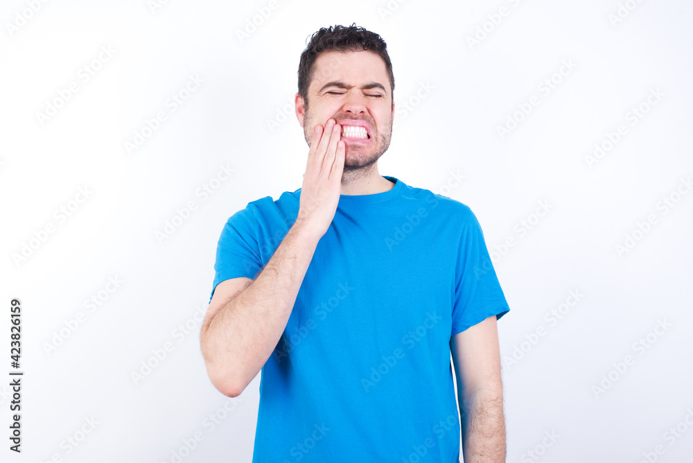 young handsome caucasian man wearing blue t-shirt against white background touching mouth with hand with painful expression because of toothache or dental illness on teeth. young handsome caucasian man wearing blue t-shirt against white background touching mouth with hand with painful expression because of toothache or dental illness on teeth.