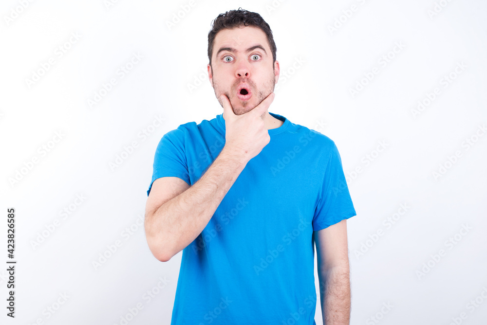 young handsome caucasian man wearing blue t-shirt against white background Looking fascinated with disbelief, surprise and amazed expression with hands on chin young handsome caucasian man wearing blue t-shirt against white background Looking fascinated with disbelief, surprise and amazed expression with hands on chin
