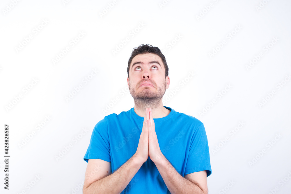 young handsome caucasian man wearing blue t-shirt against white background begging and praying with hands together with hope expression on face very emotional and worried. Please God young handsome caucasian man wearing blue t-shirt against white background begging and praying with hands together with hope expression on face very emotional and worried. Please God