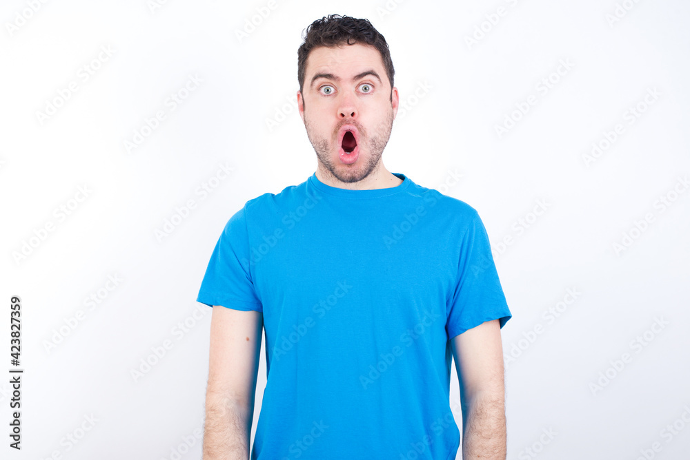 Fototapeta premium Oh my God. Surprised young handsome caucasian man wearing blue t-shirt against white background stares at camera with shocked expression exclaims with unexpectedness,