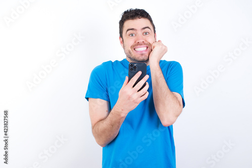 Joyous young handsome caucasian man wearing bluet-shirt poses with mobile phone device, types text message on modern smartphone, watches funny video during free time, enjoys good internet connection