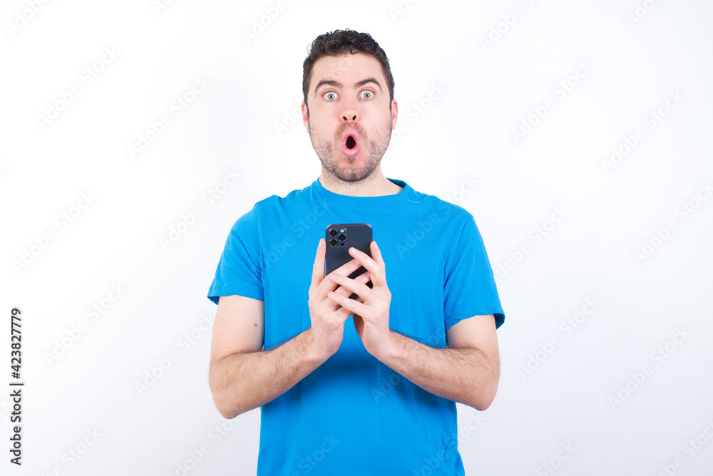 Shocked young handsome caucasian man wearing blue t-shirt against white background opens mouth hold phone reading advert unbelievable big shopping prices Shocked young handsome caucasian man wearing blue t-shirt against white background opens mouth hold phone reading advert unbelievable big shopping prices