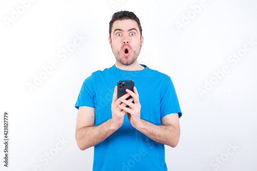 Shocked  young handsome caucasian man wearing blue t-shirt against white background opens mouth hold phone reading advert unbelievable big shopping prices © Roquillo