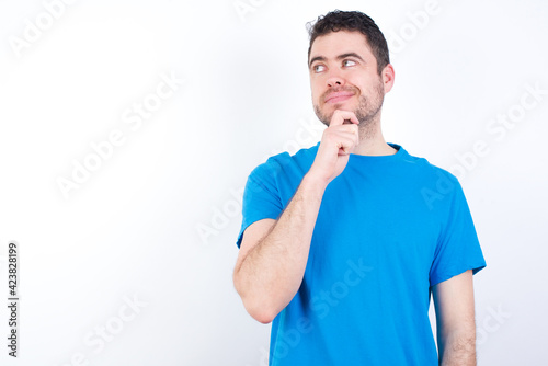 Thoughtful young handsome caucasian man wearing white t-shirt against white background holds chin and looks away pensively makes up great plan