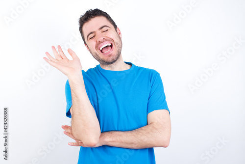 Overjoyed successful young handsome caucasian man wearing white t-shirt against white background raises palm and closes eyes in joy being entertained by friends