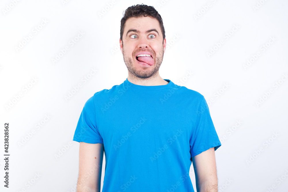 Funny young handsome caucasian man wearing white t-shirt against white background makes grimace and crosses eyes plays fool has fun alone sticks out tongue. Funny young handsome caucasian man wearing white t-shirt against white background makes grimace and crosses eyes plays fool has fun alone sticks out tongue.