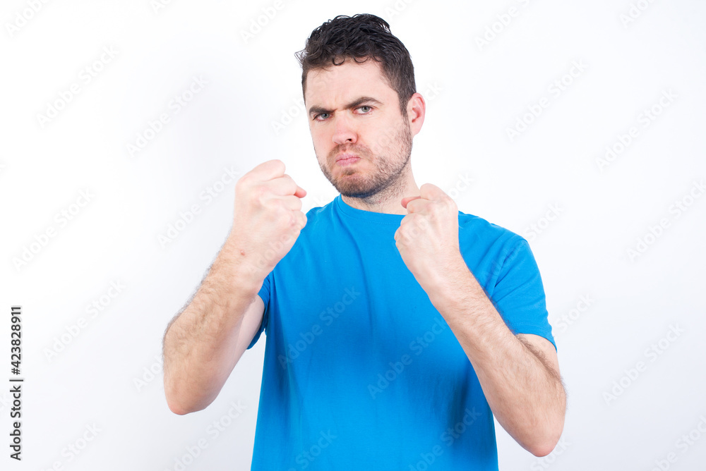 Displeased annoyed young handsome caucasian man wearing white t-shirt clenches fists, gestures pissed, ready to revenge, looks with aggression at camera stands full of hate, being pressured Displeased annoyed young handsome caucasian man wearing white t-shirt clenches fists, gestures pissed, ready to revenge, looks with aggression at camera stands full of hate, being pressured