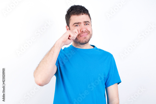 Disappointed dejected young handsome caucasian man wearing white t-shirt against white background wipes tears stands stressed with gloomy expression. Negative emotion