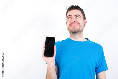 young handsome caucasian man wearing white t-shirt against white background holds new mobile phone and looks mysterious aside shows blank display of modern cellular