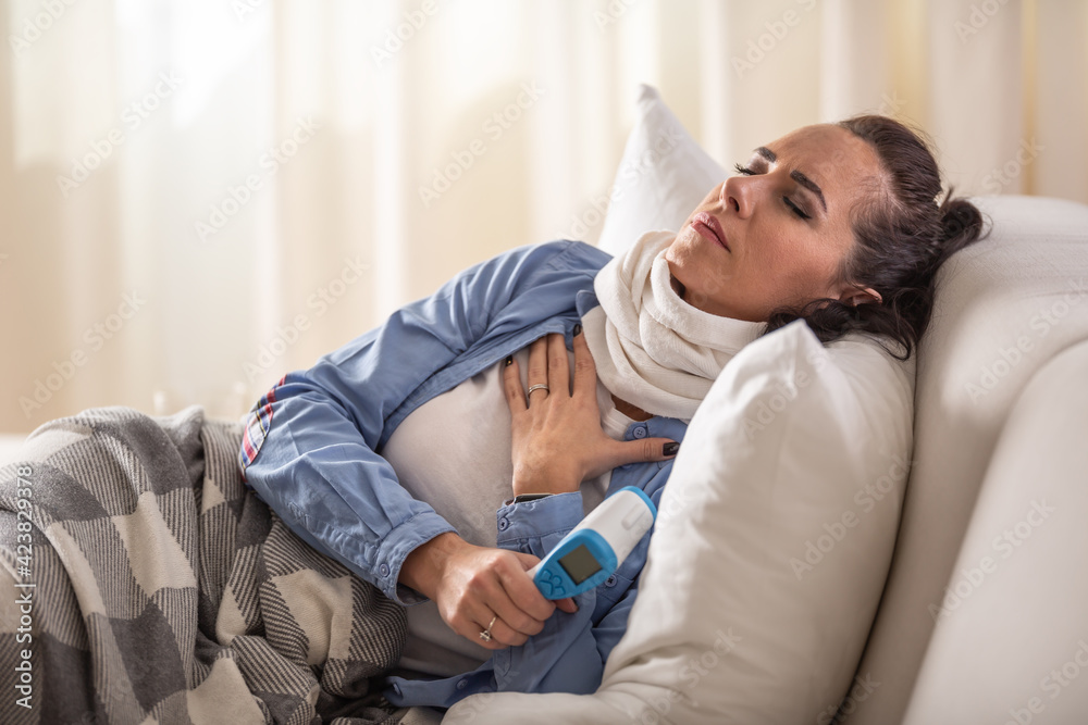 Fototapeta premium Ill woman experiencing breathing and cough problems while holding forehhead thermometer and lying down on a sofa