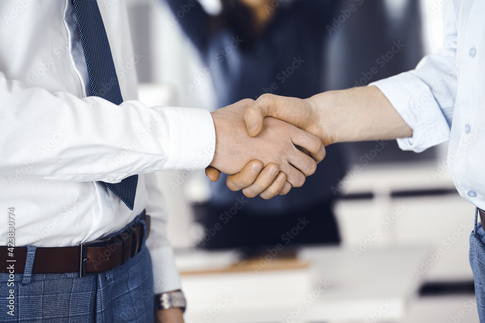 Fototapeta premium Two businessmen are shaking hands in office, close-up. Happy and excited business woman stands with raising hands at the background. Business people concept