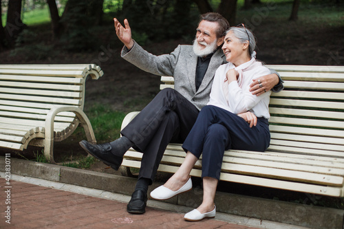 Stylish elderly couple sitting in embrace on wooden bench. Happy mature man and woman talking during date at green park.