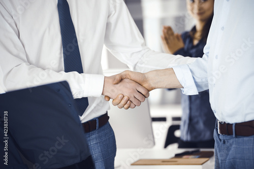 Two businessmen are shaking hands in office  close-up. Happy and excited business woman stands with raising hands at the background. Business people concept