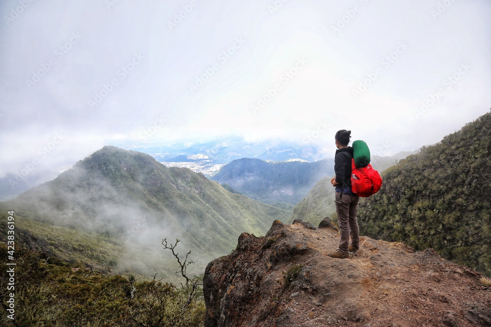 Senderista latinoamericana con mochila color rojo desde una roca mirando hacia montañas y paisaje con nubes lleno de naturaleza