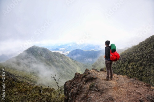 Senderista latinoamericana con mochila color rojo desde una roca mirando hacia montañas y paisaje con nubes lleno de naturaleza © AALM