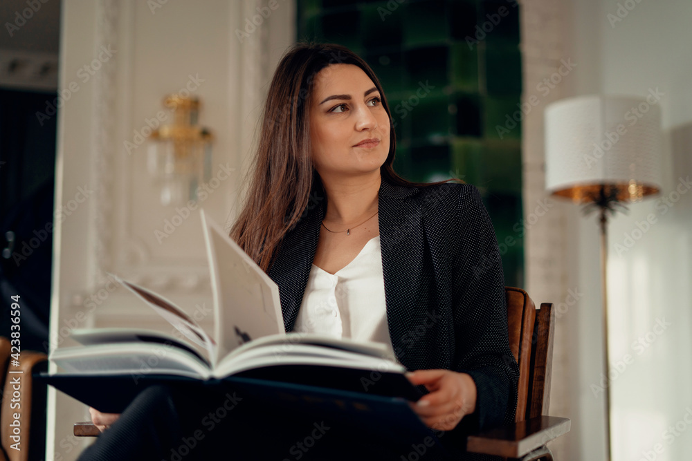 A satisfied finance manager. He is sitting on a chair, holding a book with work reports in his hands. A businesswoman is in a modern, stylish office. Clothing business suit in black. A satisfied finance manager. He is sitting on a chair, holding a book with work reports in his hands. A businesswoman is in a modern, stylish office. Clothing business suit in black.