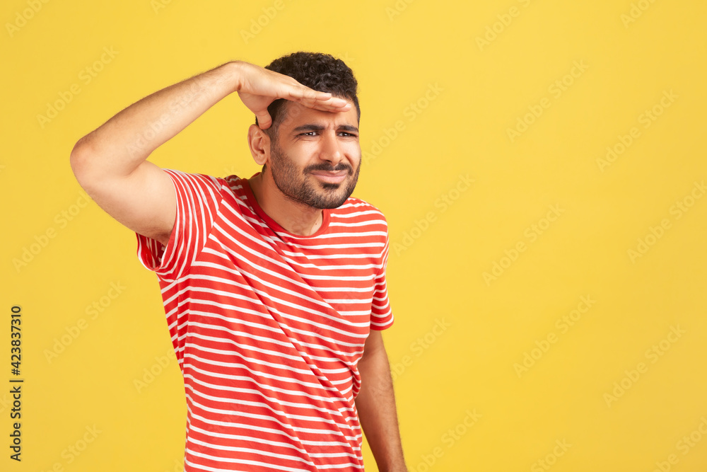 Fototapeta premium Nosy attentive man with beard in red striped t-shirt looking far away holding hand near forehead, watching at distance, searching for something. Indoor studio shot isolated on yellow background